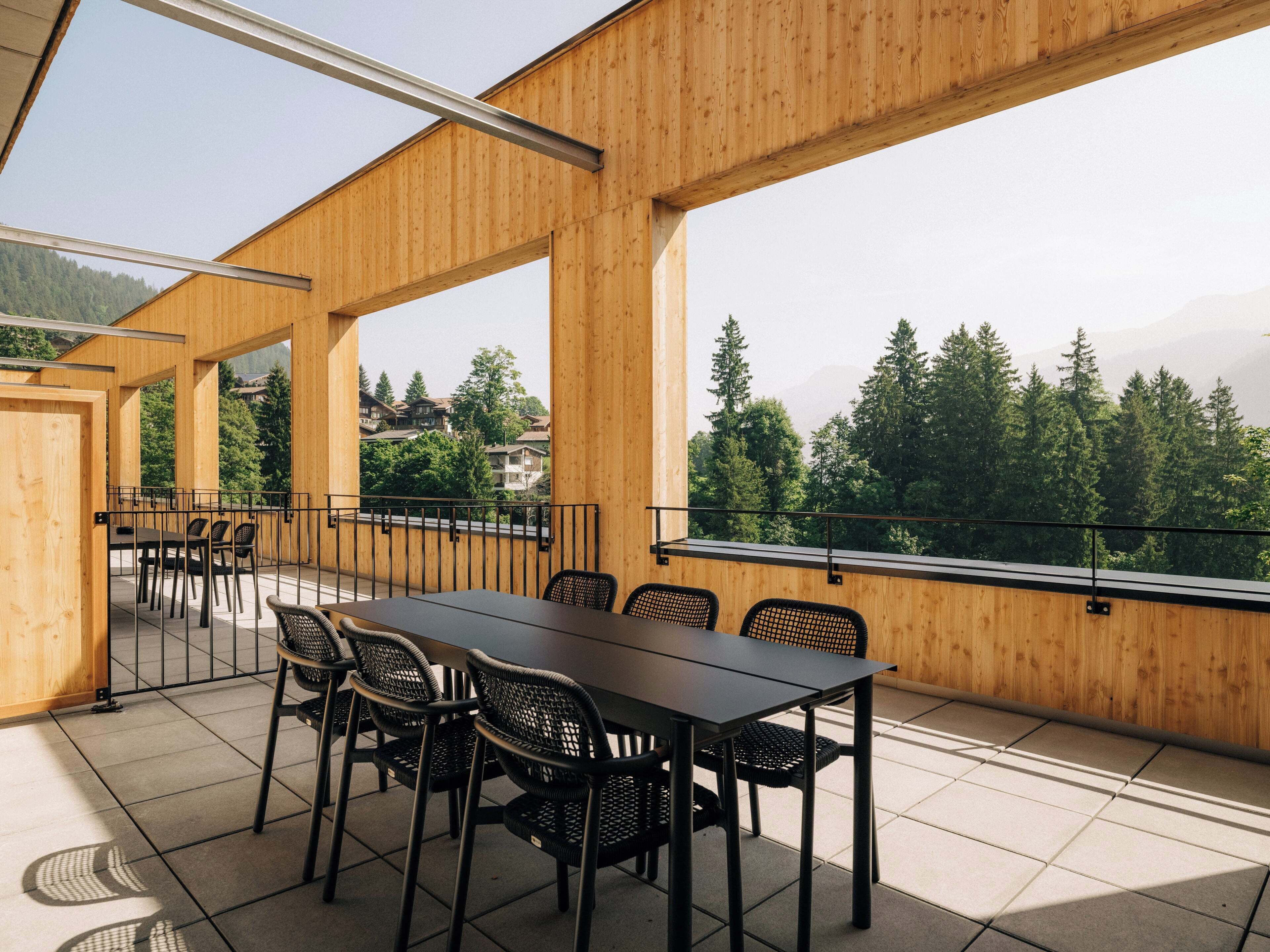 A wooden deck with a table and chairs, large windows offering a view of trees and mountains.