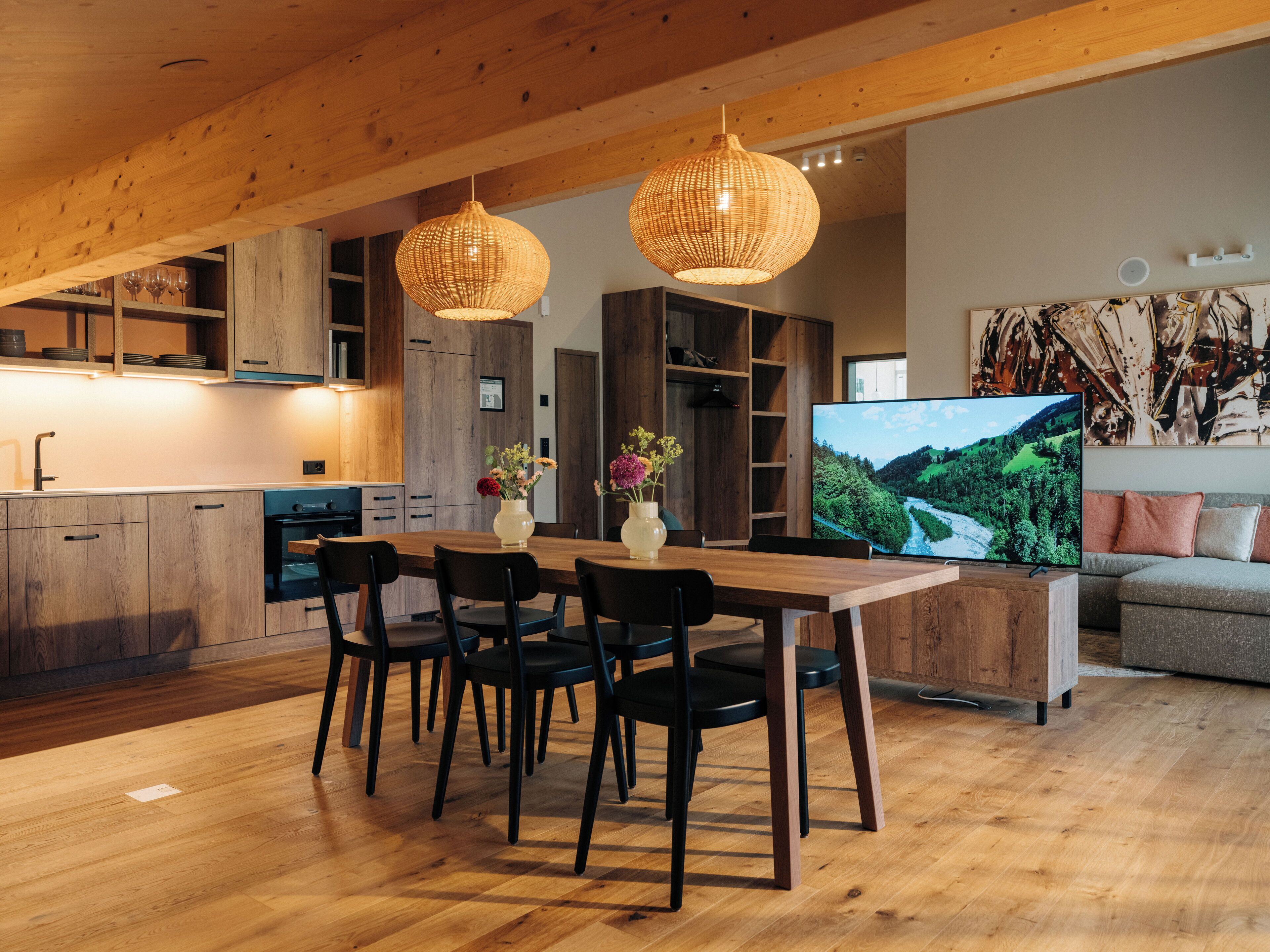 A modern kitchen with wooden cabinets, a dining table with chairs, and a large flat-screen TV displaying a scenic landscape.