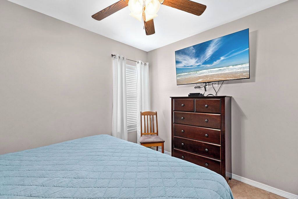 A bedroom with a bed, a dresser, a chair, a ceiling fan, and a beach scene on the wall.