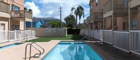 A swimming pool surrounded by apartment buildings, with a fenced area and a grassy patch in the background.