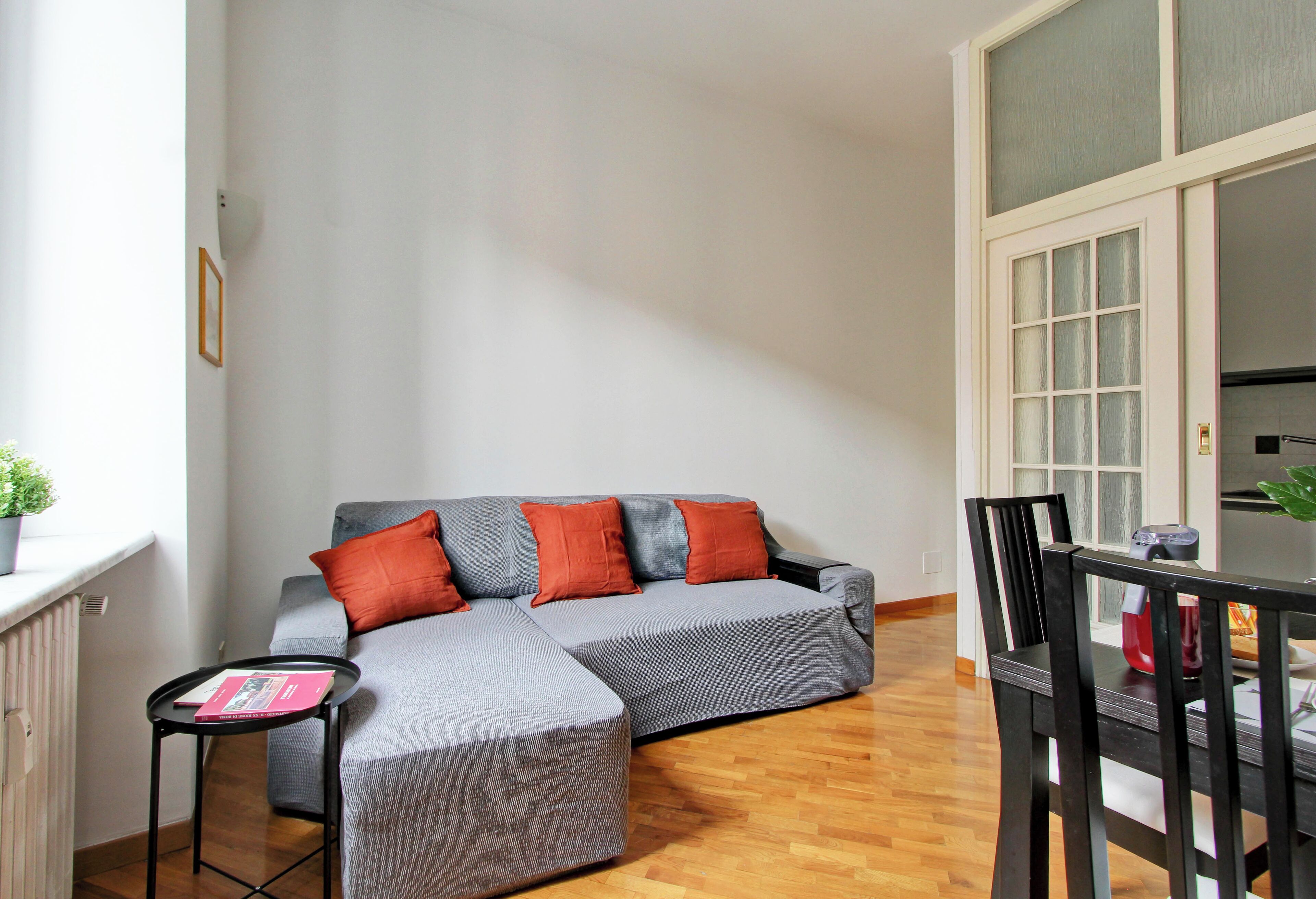 A modern living room with a grey sectional sofa, red cushions, a small black side table, and a dining area with a black chair and table.
