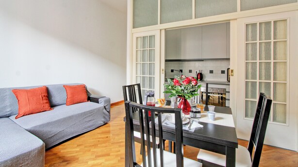 Ein modernes Wohnzimmer mit grauem Sofa, roten Kissen, einem Esstisch mit Stühlen und einer Küche im Hintergrund.