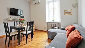 Ein modernes Wohnzimmer mit Flachbildfernseher, Esstisch mit Stühlen, grauem Sofa und orangefarbenen Kissen sowie einem Fenster mit Heizkörper.