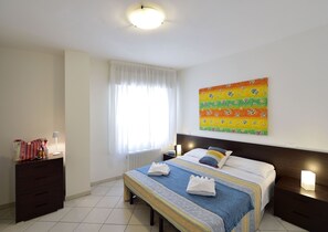 A hotel room with a bed, bedside tables, a nightstand, a window with curtains, a radiator, a wall art piece, and a lamp. - Mina Suite - Double Apartment (Cattolica)