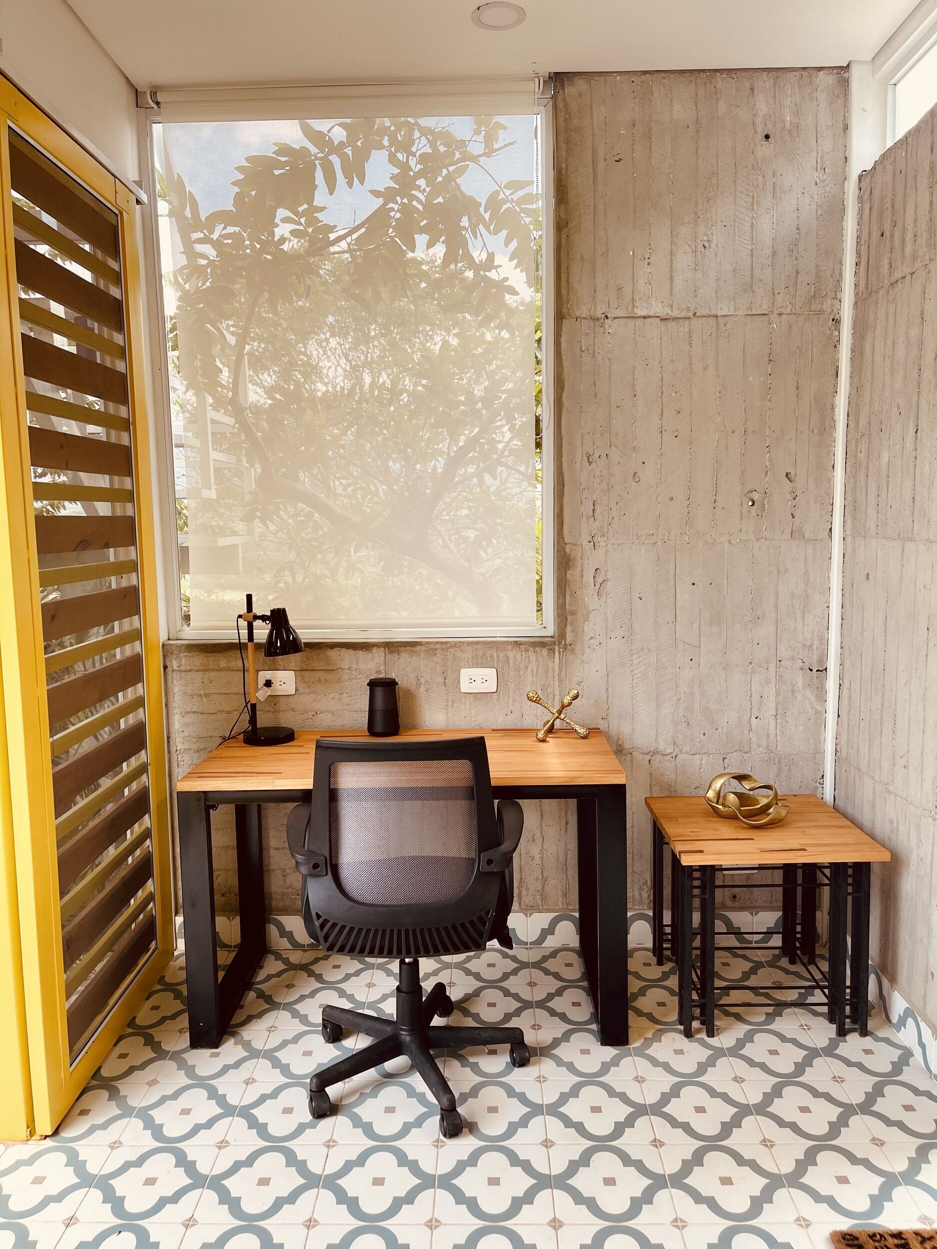 Un petit bureau avec un bureau en bois, une chaise de bureau noire, une petite table d’appoint et une fenêtre donnant sur des arbres.
