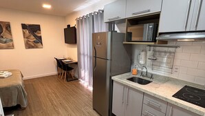 Een compacte keuken met een koelkast, magnetron en spoelbak. Er is een eethoek met een tafel en stoelen, en een klein raam met gordijnen.