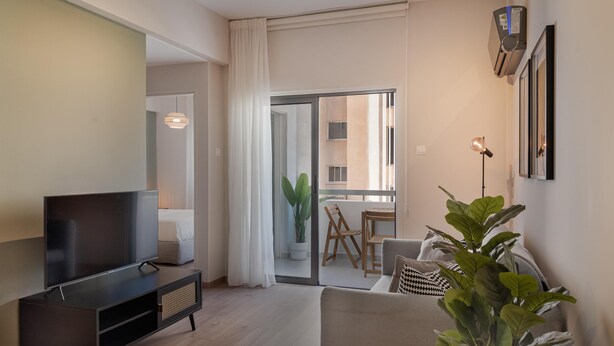 Una sala moderna con un televisor de pantalla plana sobre un soporte negro, un sofá gris, una planta en maceta y un balcón con una mesa y sillas de comedor.