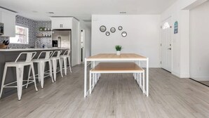 Moderni keittiö, jossa on valkoiset kaapit, keskellä sijaitseva saareke baarituoleineen sekä ruokapöytä, jonka päällä on ruukkukasvi.