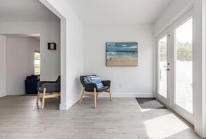 A modern living room with a large window, a framed beach painting, and a dark-colored sofa.