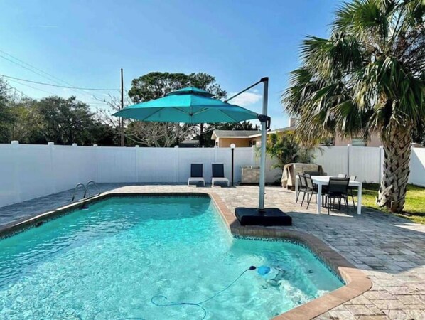 A rectangular swimming pool with a tiled edge, a lounge chair, and a table with chairs in the background.