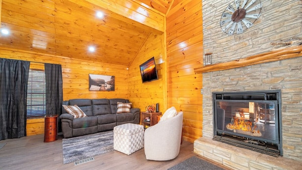 A cozy log cabin interior with a stone fireplace, wooden walls, a sofa, and a television mounted on the wall. - *Creekside 1BD + Loft Cabin! Hot Tub + Fire Pit!* (Sevierville)