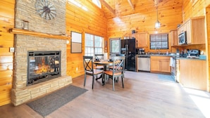 A log cabin interior with a stone fireplace, wooden dining table, chairs, kitchen area, and a clock on the wall. - *Creekside 1BD + Loft Cabin! Hot Tub + Fire Pit!* (Sevierville)