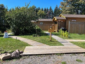 A wooden cabin with a deck, surrounded by a grassy yard and trees.