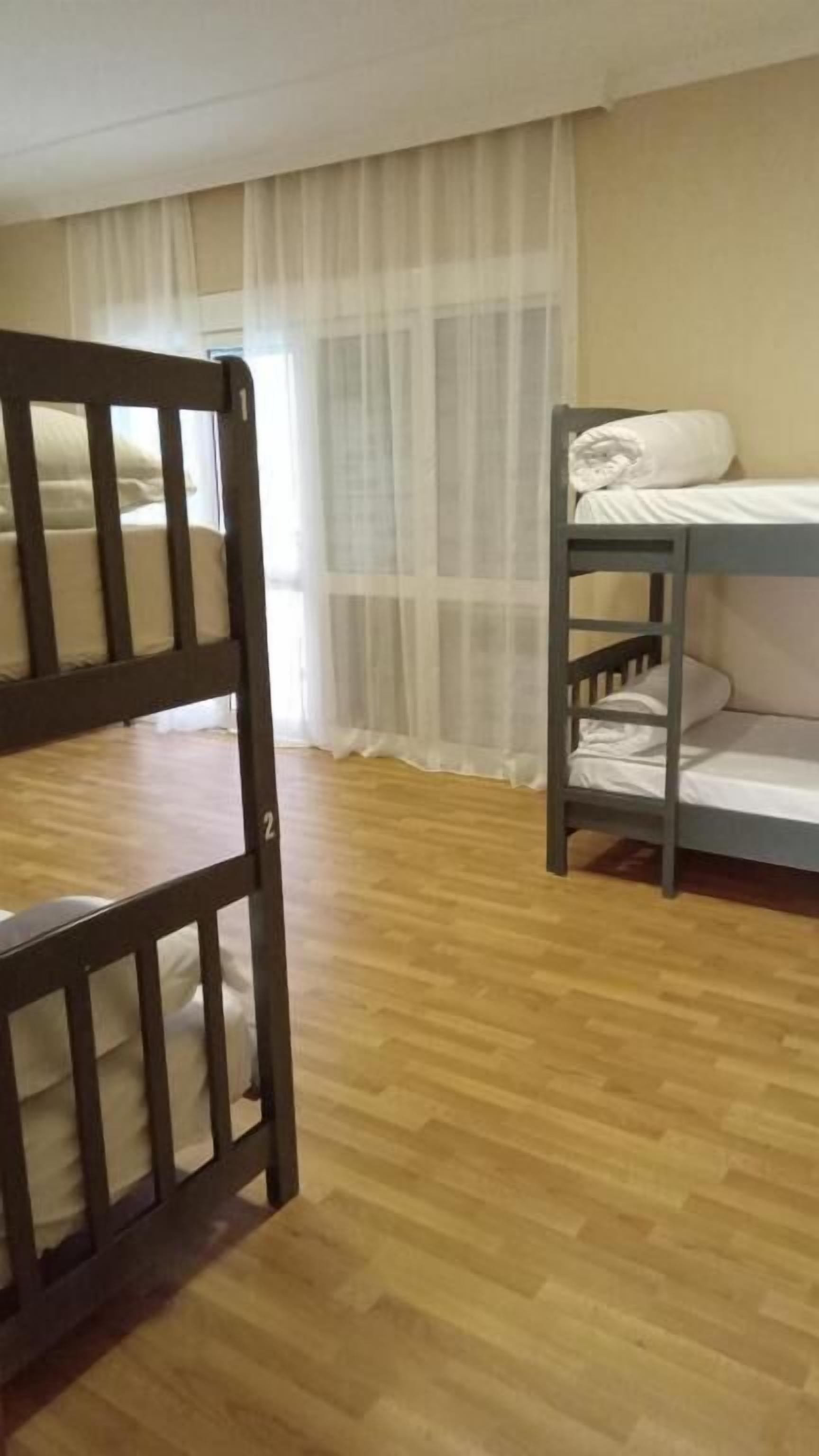A room with two bunk beds, a wooden floor, and a sheer curtain.