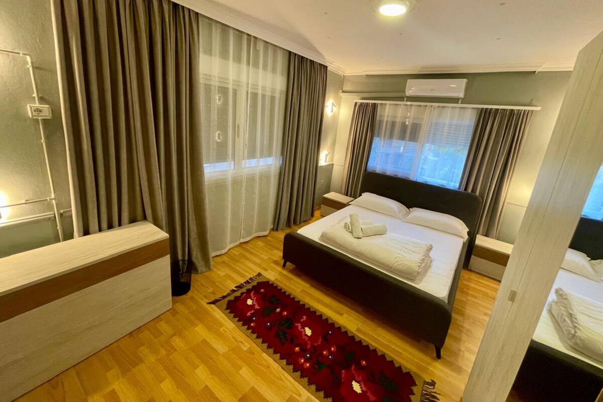 A hotel room with a bed, a bedside table, a desk, a window with curtains, an air conditioning unit, and a red decorative rug.