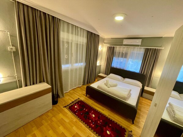 Habitación de hotel con cama, mesita de noche, escritorio, ventana con cortinas, equipo de aire acondicionado y una alfombra decorativa roja.