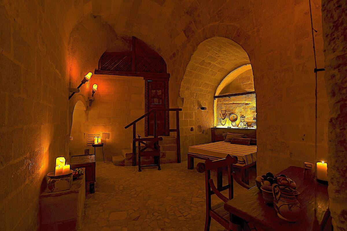 A dimly lit room with stone walls, a wooden bed, a bench, and a table with candles.