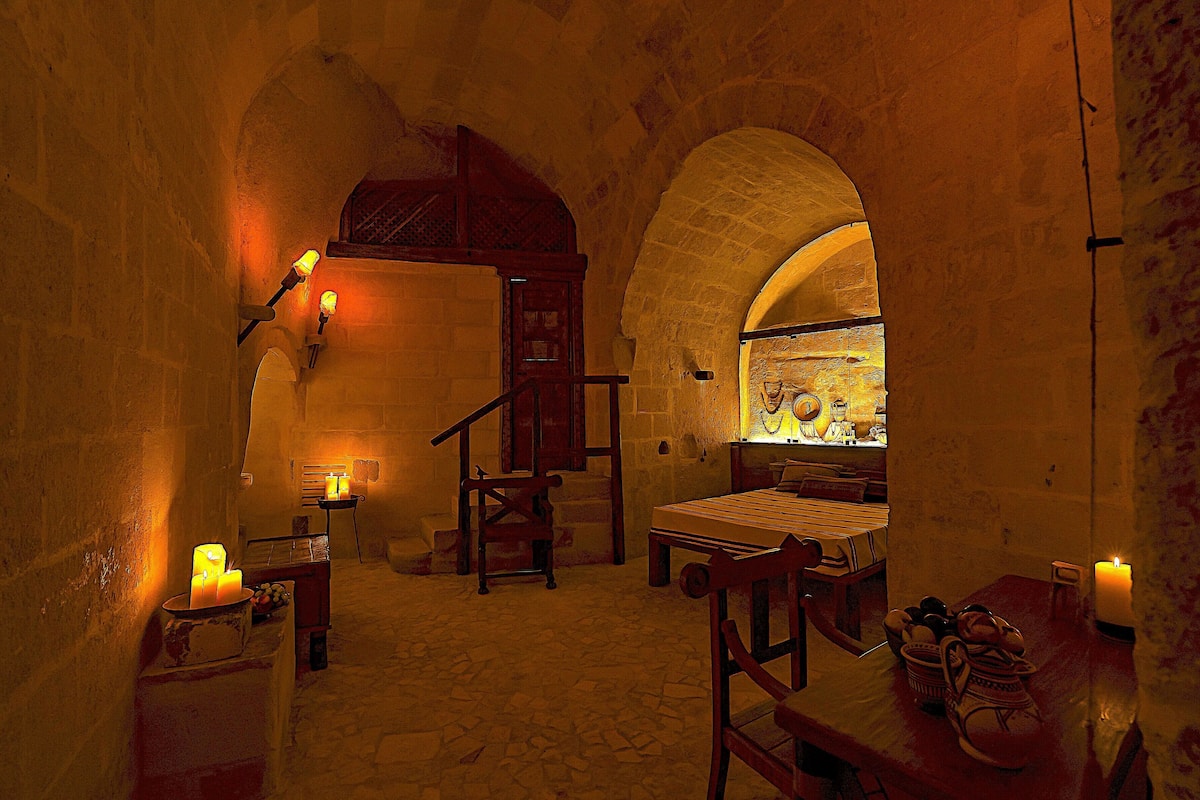 A dimly lit room with stone walls, a wooden bed, a bench, and a table with candles.