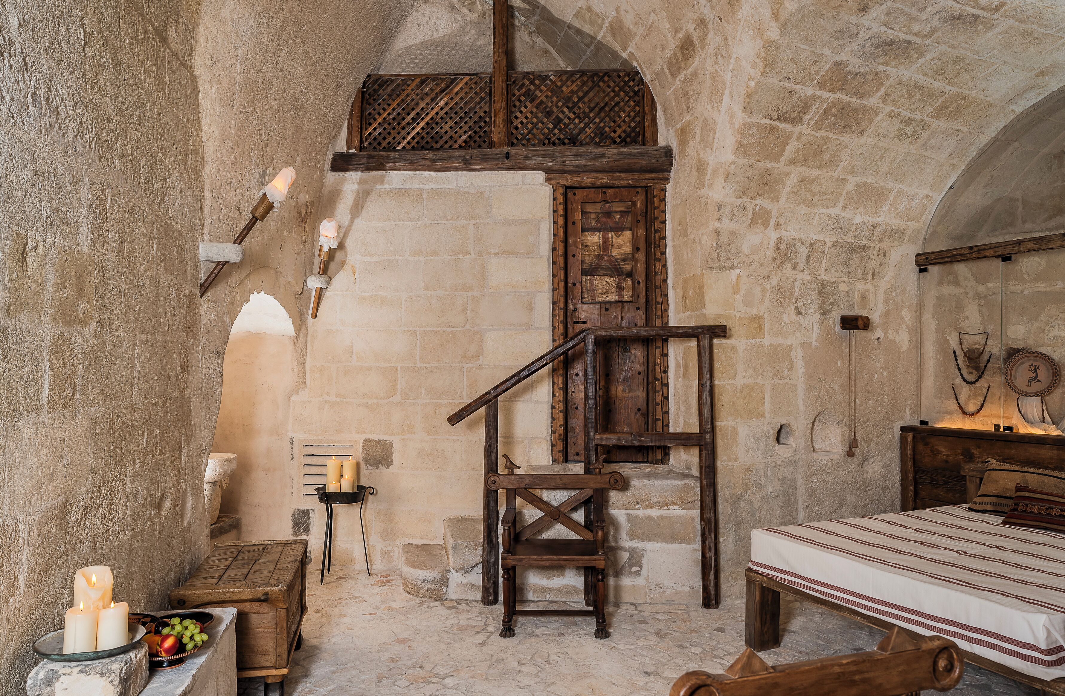A rustic room with a stone wall, wooden bed, a wooden chair, a small table with candles, and a ladder.