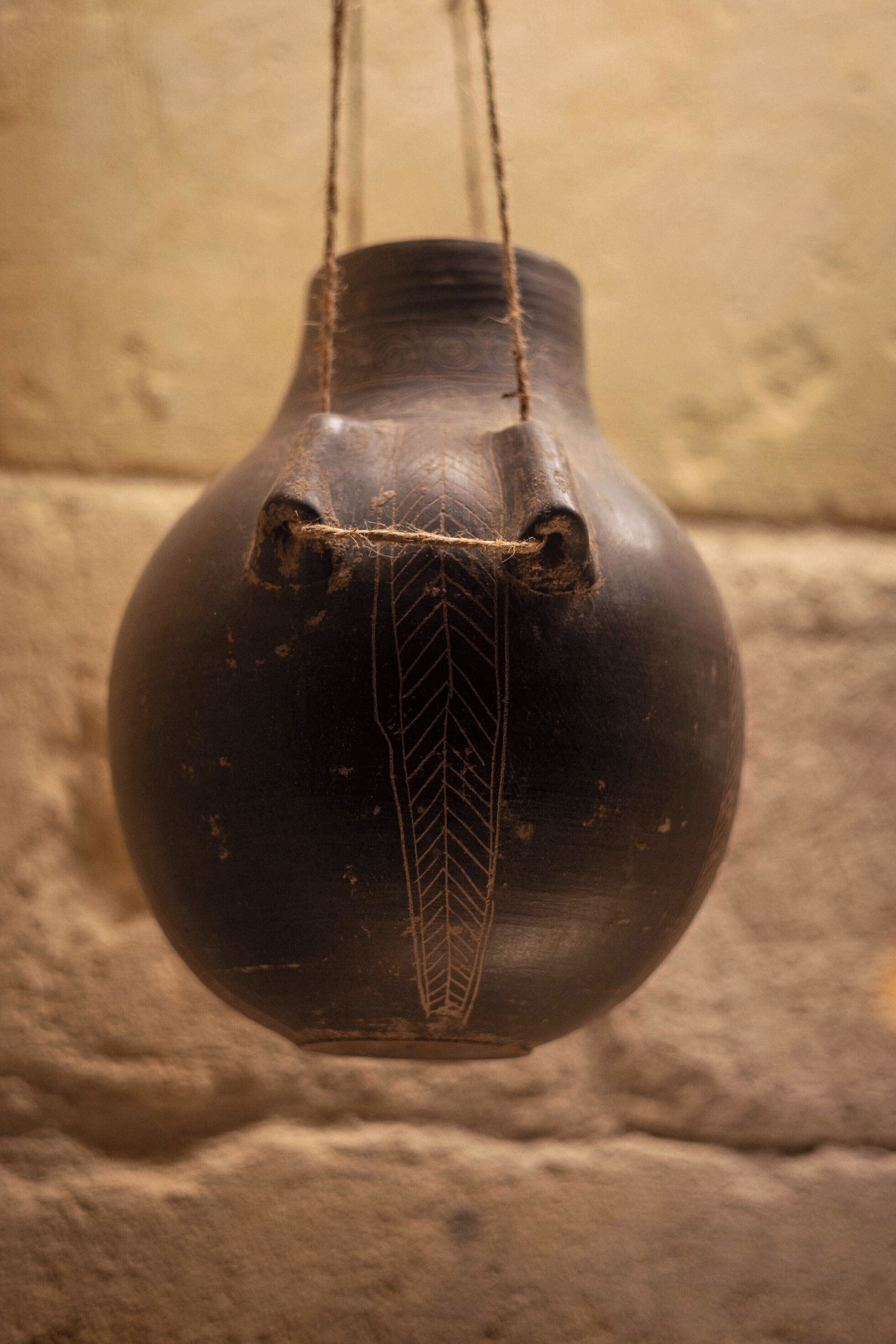 Un jarrón de cerámica oscuro, colgante y con diseño de hojas, sostenido por cuerdas.