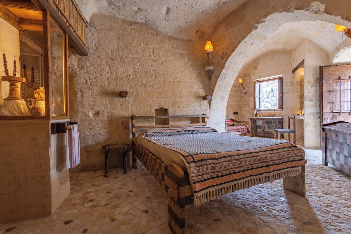 A rustic room with a stone wall, a bed with a patterned blanket, a small table, and a window with a view.