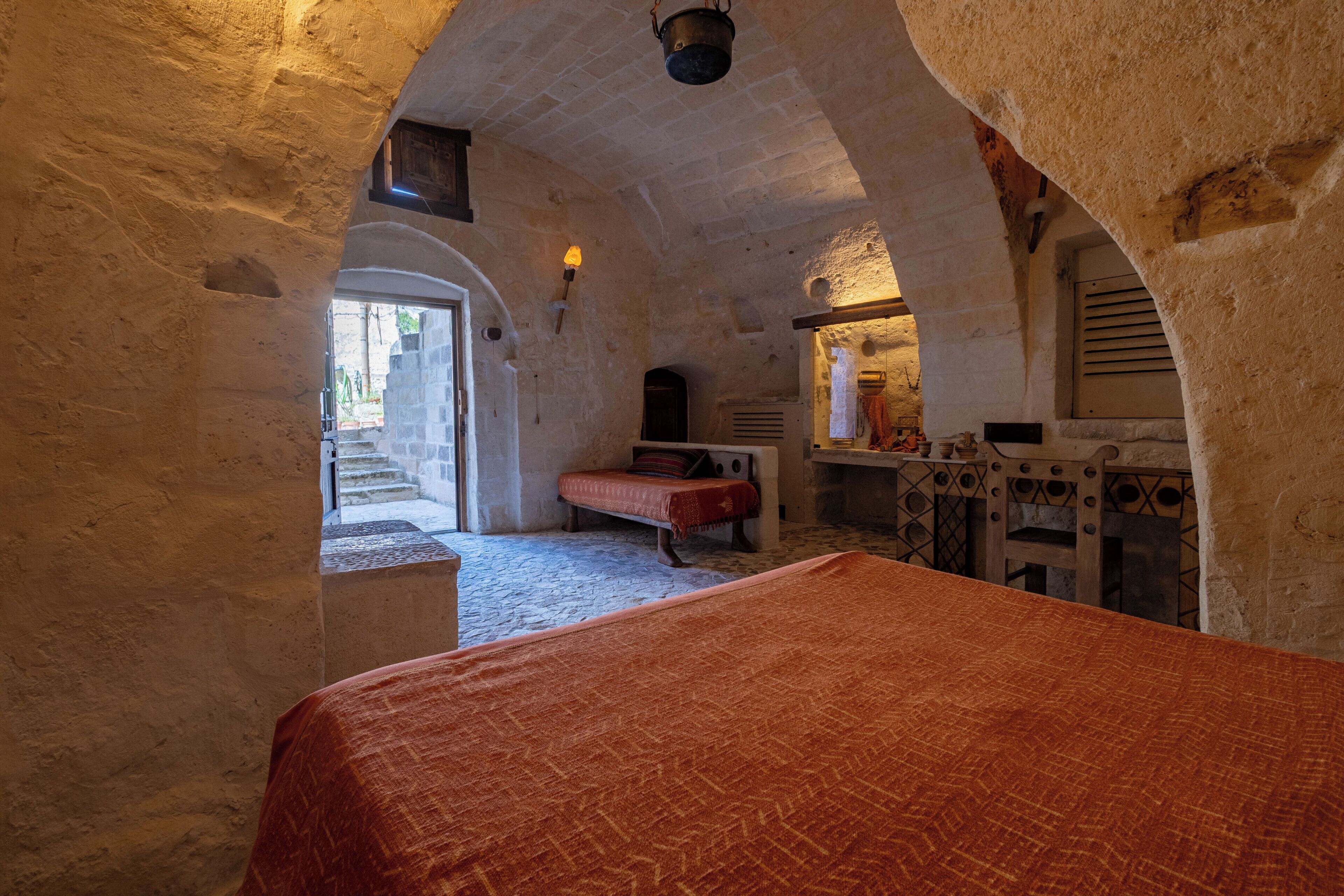 A rustic stone room with a bed, a bench, and a small table with a lamp.