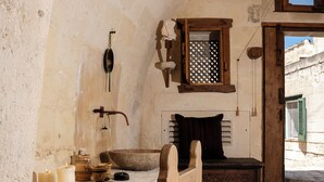Una habitación rústica con pared de piedra, un banco de madera, una pequeña mesa con velas y una ventana con contraventanas verdes.