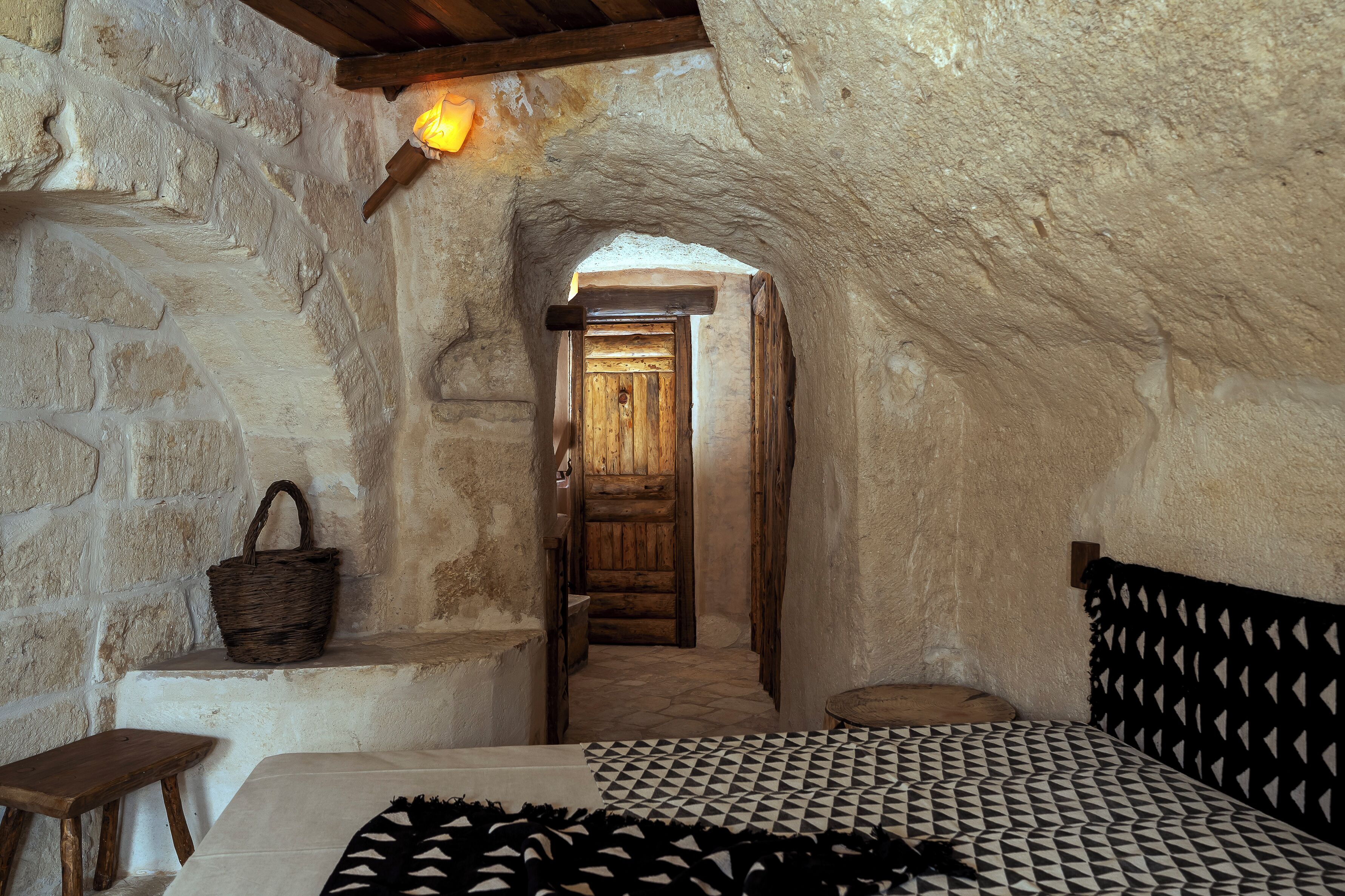 A rustic stone-walled room with a wooden door, a woven basket, a small stool, and a patterned bedspread.