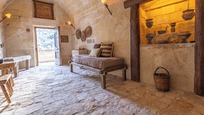 Una habitación rústica con piso de piedra, una cama con almohadas, un banco de madera, una mesa y una cesta.