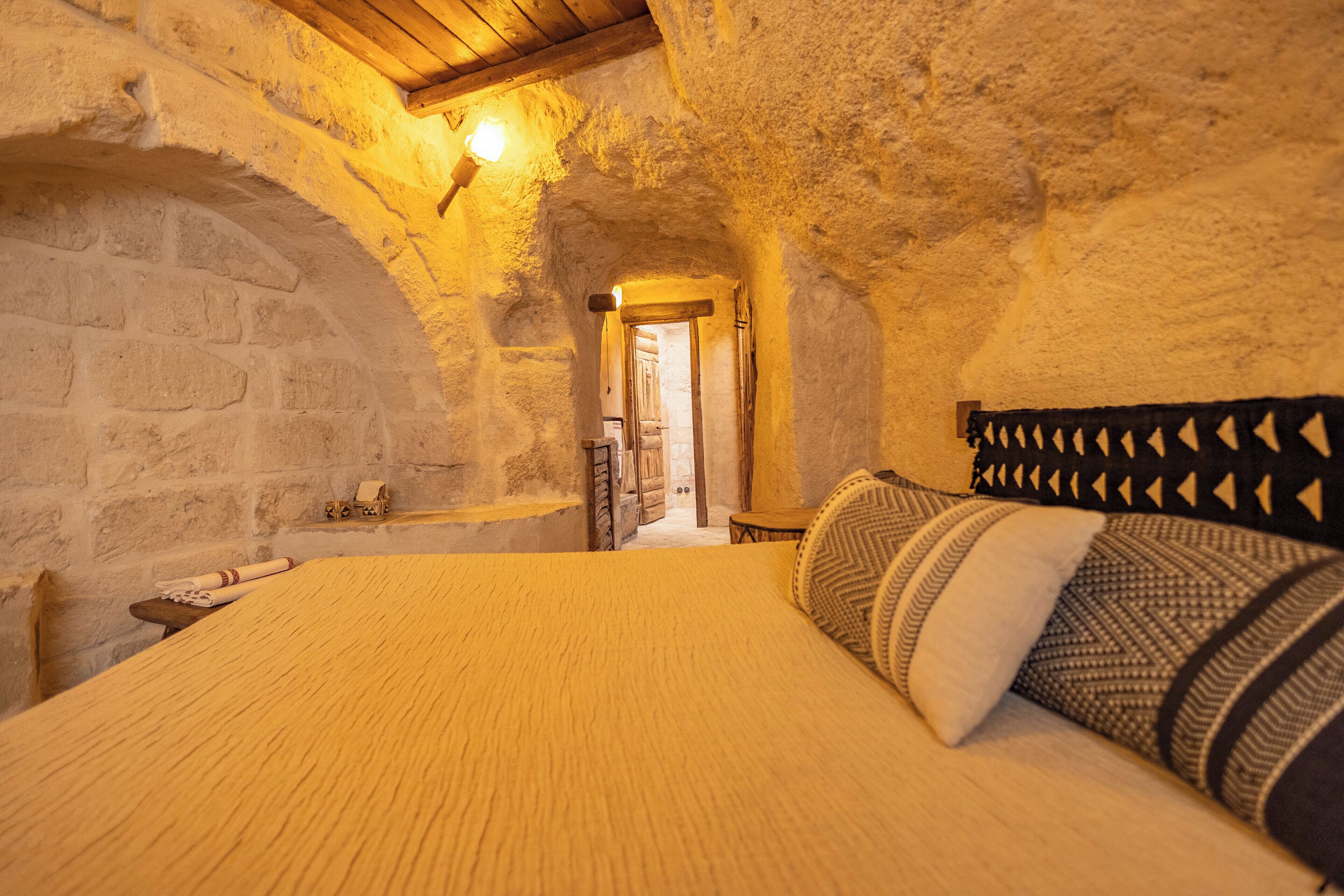 A bedroom with a stone wall, a wooden ceiling, a bed with patterned pillows, and a nightstand.