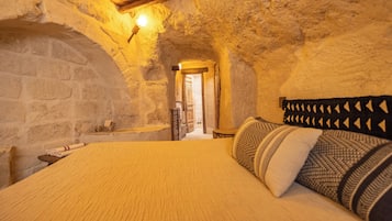 A bedroom with a stone wall, a wooden ceiling, a bed with patterned pillows, and a nightstand.