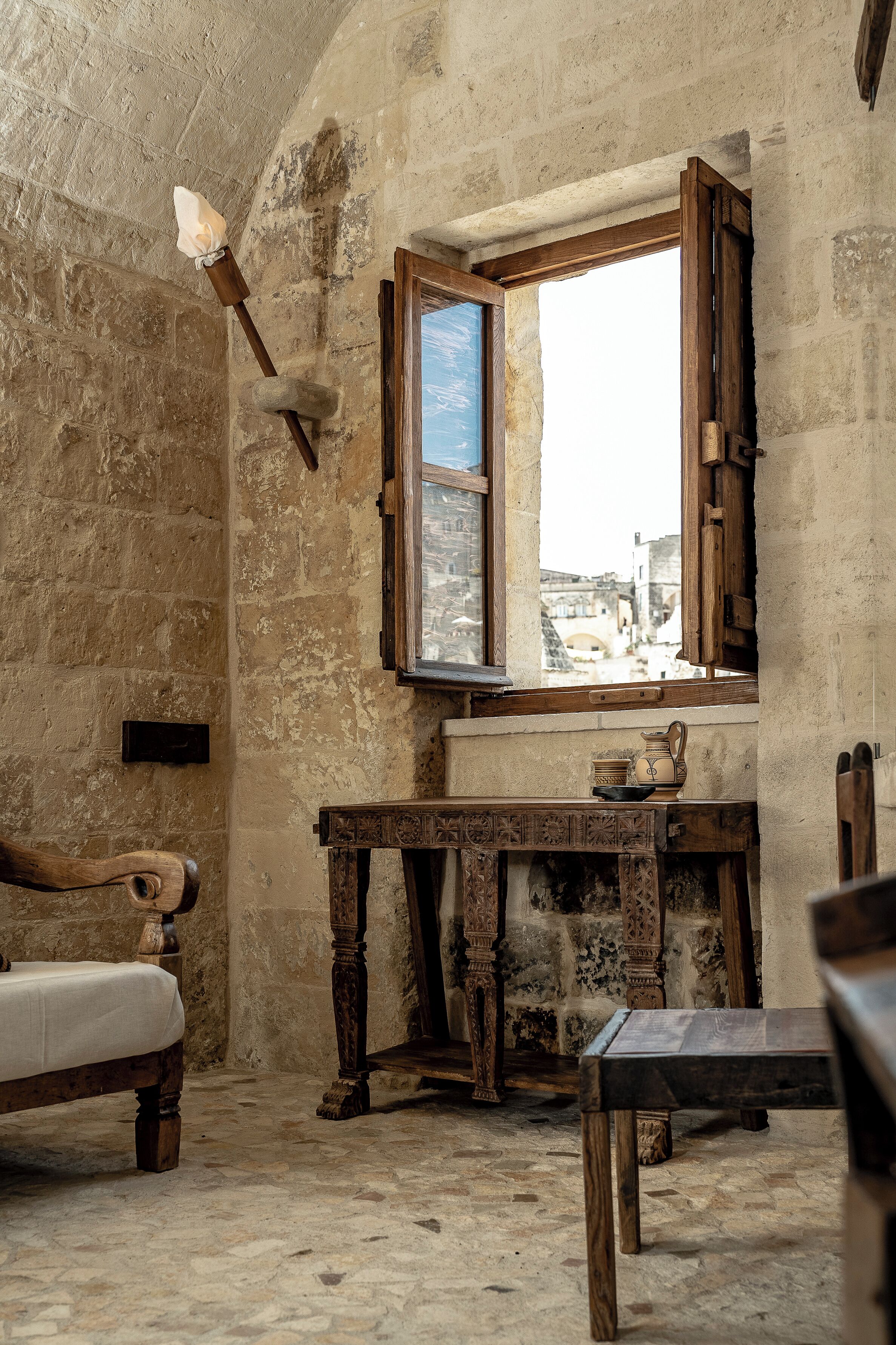 An old room with a stone wall, a wooden bed, a wooden table, and a window with a view of a bridge.