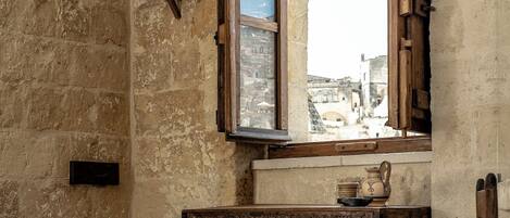 An old room with a stone wall, a wooden bed, a wooden table, and a window with a view of a bridge.