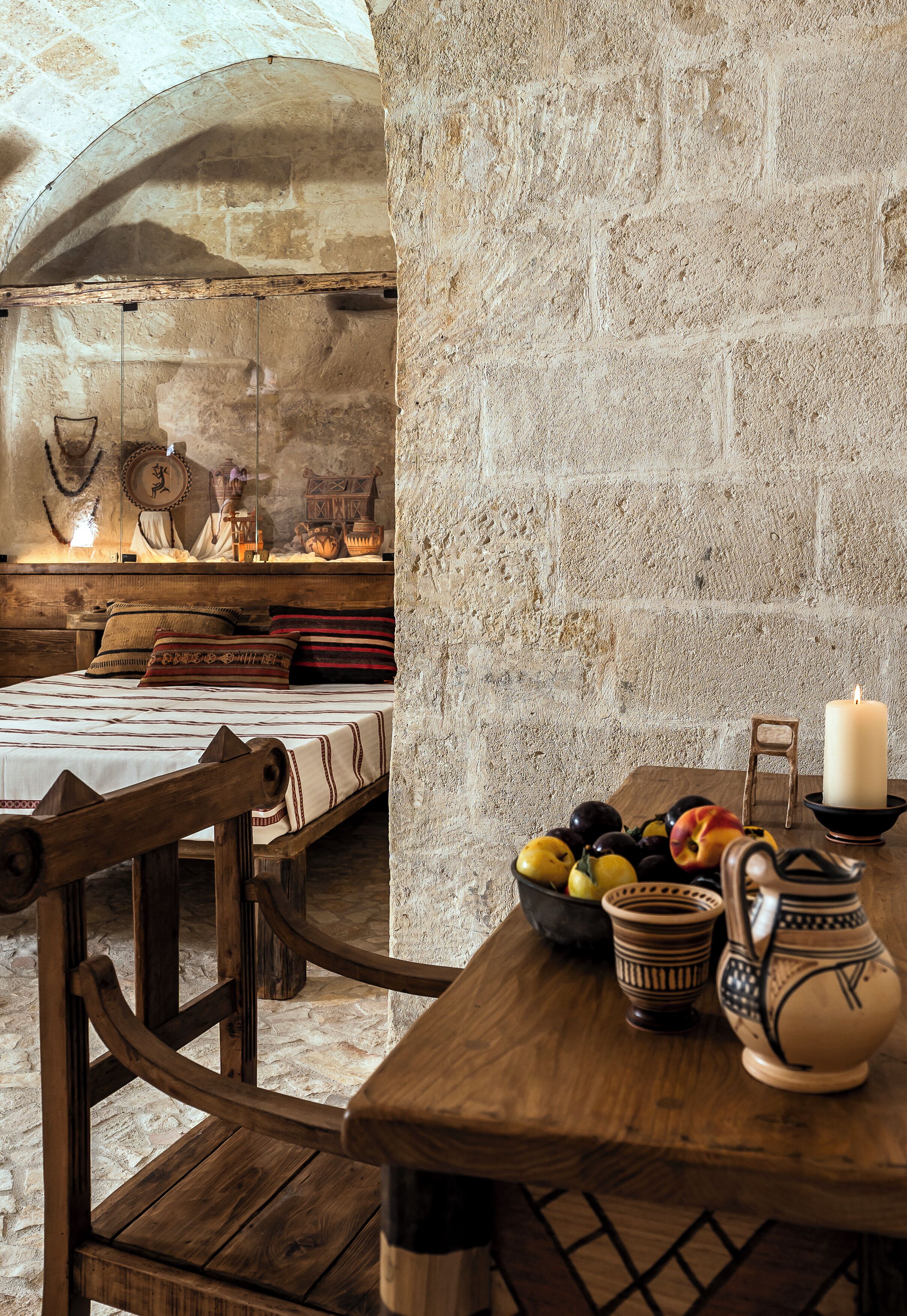 A rustic room with a wooden bed, a table with fruits, pottery, and a candle.