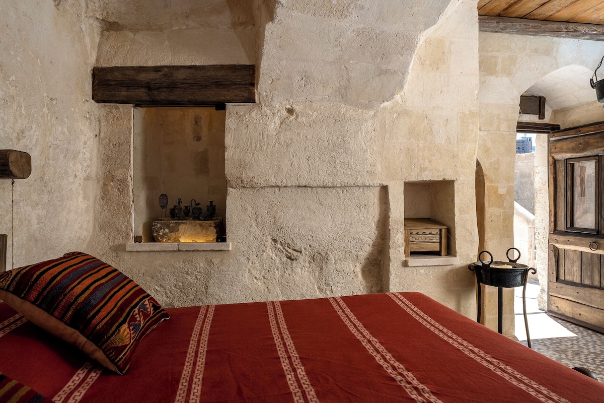 A rustic room with a bed, a shelf with bottles, a wooden door, and a stone wall.