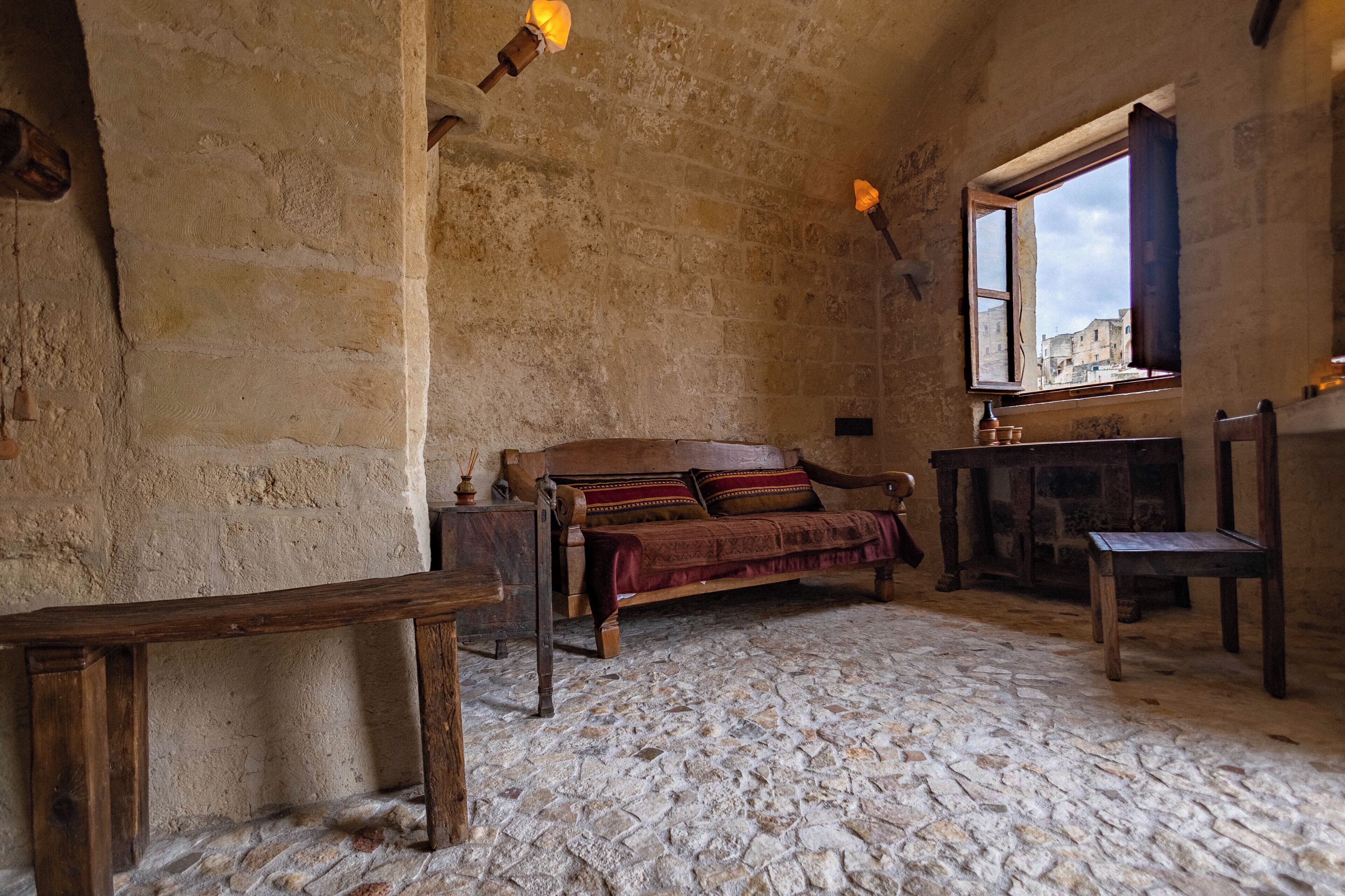 Una habitación rústica con piso de piedra, un banco de madera, una cama de madera con una manta roja, una mesita y una ventana con vista a edificios.