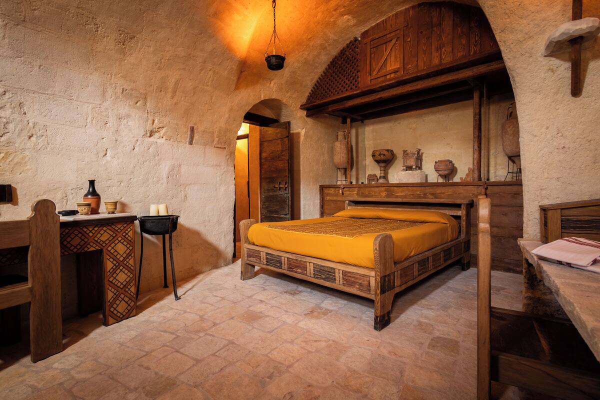 A rustic room with a wooden bed, a stone floor, and a wooden desk with pottery.