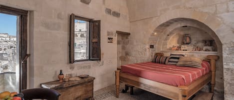 A room with a wooden bed, a wooden nightstand, a bowl of fruit, and a view of the city through a window.