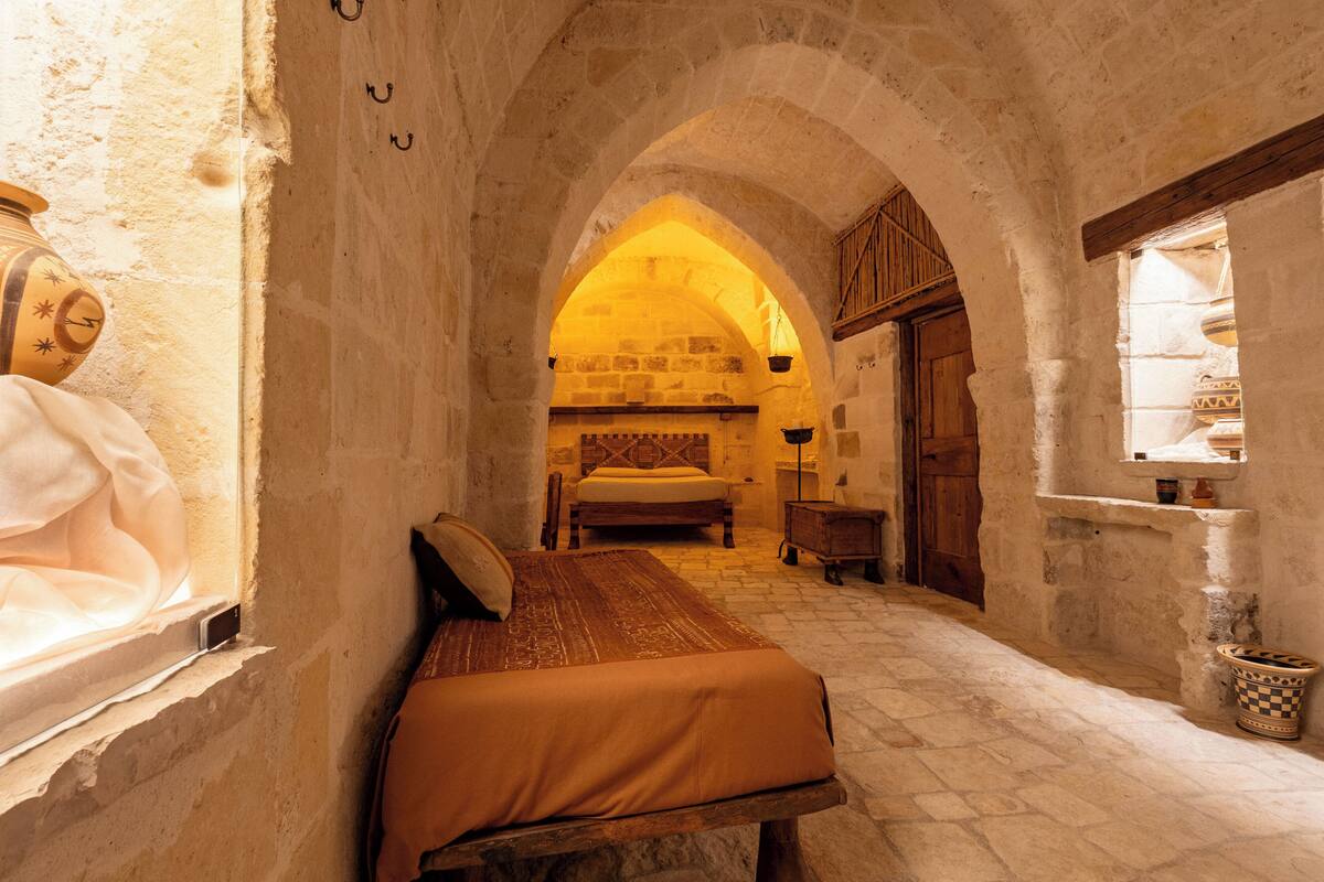 A narrow corridor with a bed, a wooden bench, and a window with a view of a room with a bed and a wooden cabinet.