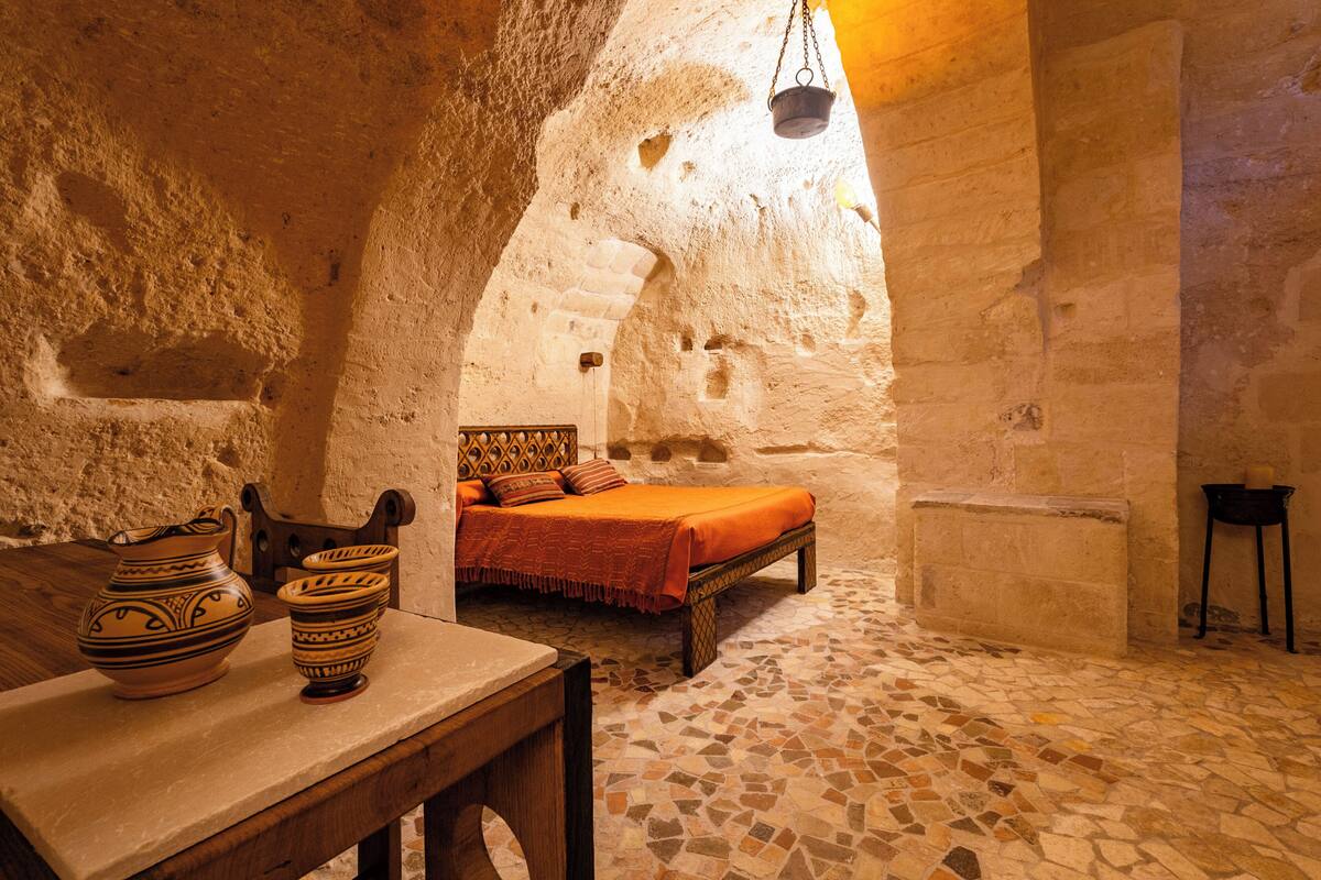 A stone-walled room with a bed, a table with pottery, and a hanging pot.