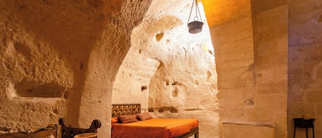 Una habitación con paredes de piedra, una cama, una mesa con cerámica y una olla colgante.