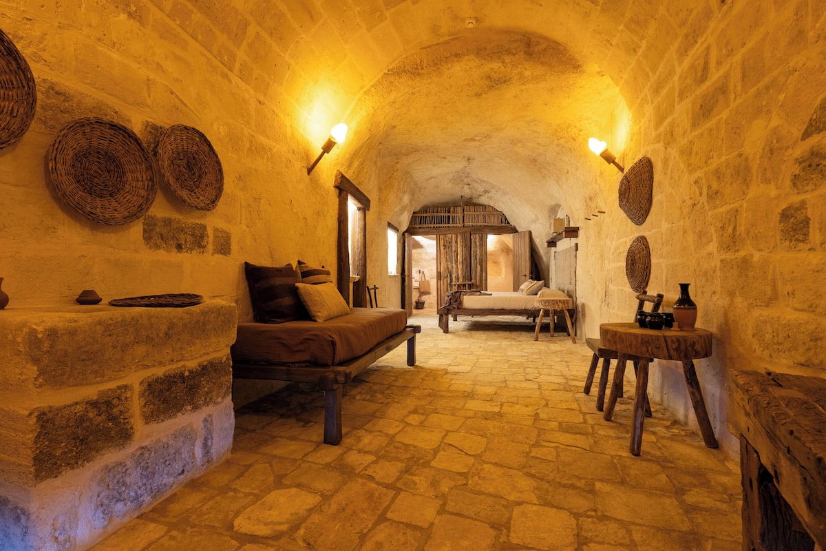 A stone-walled room with a bed, a bench, and a table with a vase.