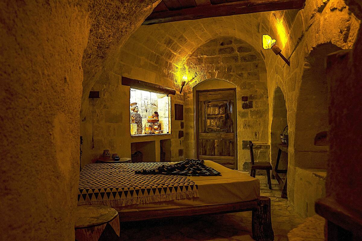 A room with a bed, a window with a view, a wooden door, and a stone wall.