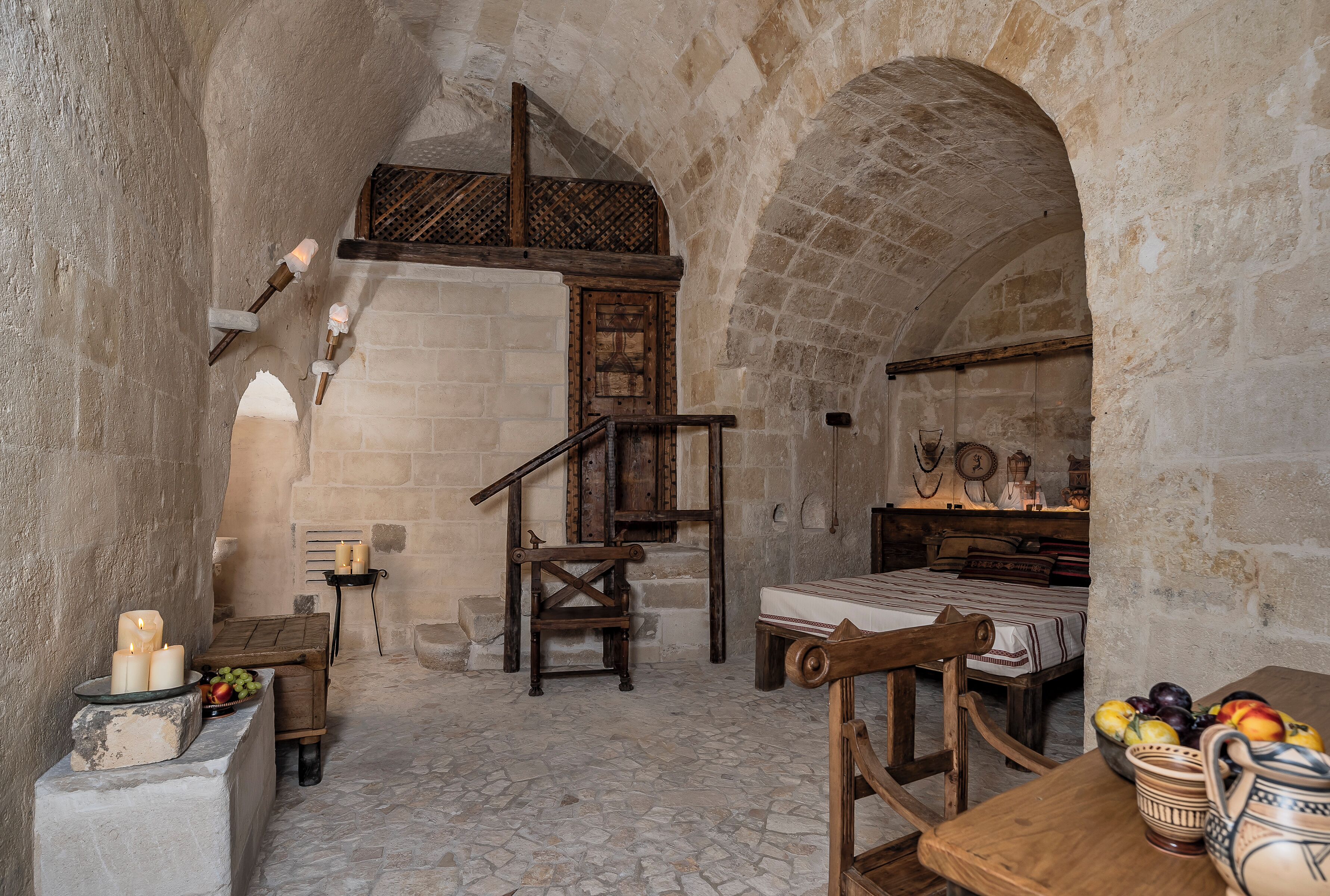 A stone-walled room with a bed, a wooden chair, a table with fruit, and a shelf with candles.