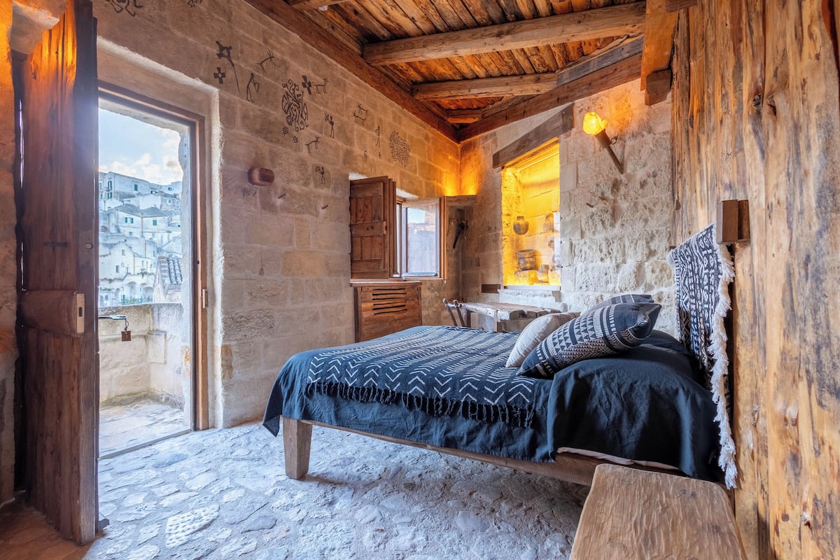 A rustic bedroom with a stone floor, wooden beams, a bed with patterned bedding, a wooden desk, and a view of a town through a doorway.