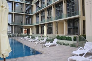A pool area with lounge chairs and umbrellas, surrounded by a building with multiple floors and large windows.