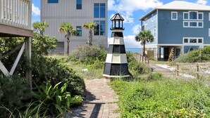 Un sendero conduce a un edificio blanco con un faro, rodeado de follaje y una terraza de madera.