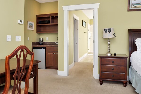 A hotel room with a wooden chair, a bedside table with a lamp, a bed, a nightstand, a dresser, a kitchen area, and a hallway leading to another room. - Adventure Time (Port St. Joe)