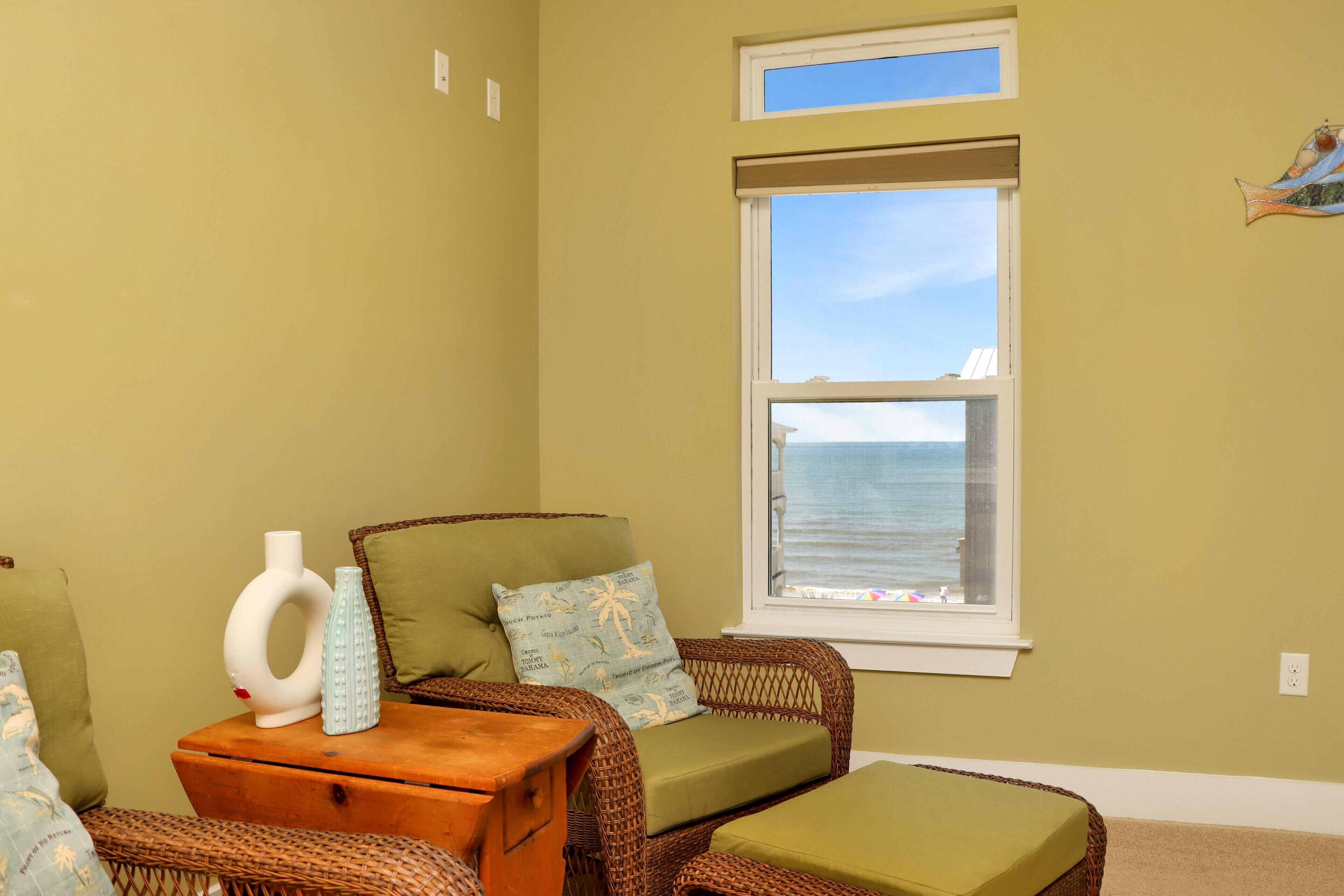 A room with a wicker chair, a small wooden table, and a window with a view of the ocean.