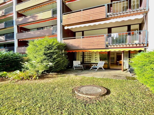 A multi-story residential building with balconies, a small patio area with two chairs, and a manicured lawn.